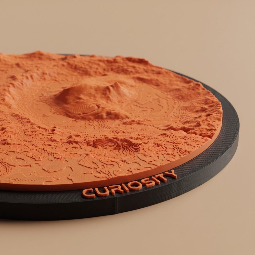 A low-angle close-up of the Curiosity ornament: the layered relief of Mount Sharp in red, with raised CURIOSITY lettering on the black frame