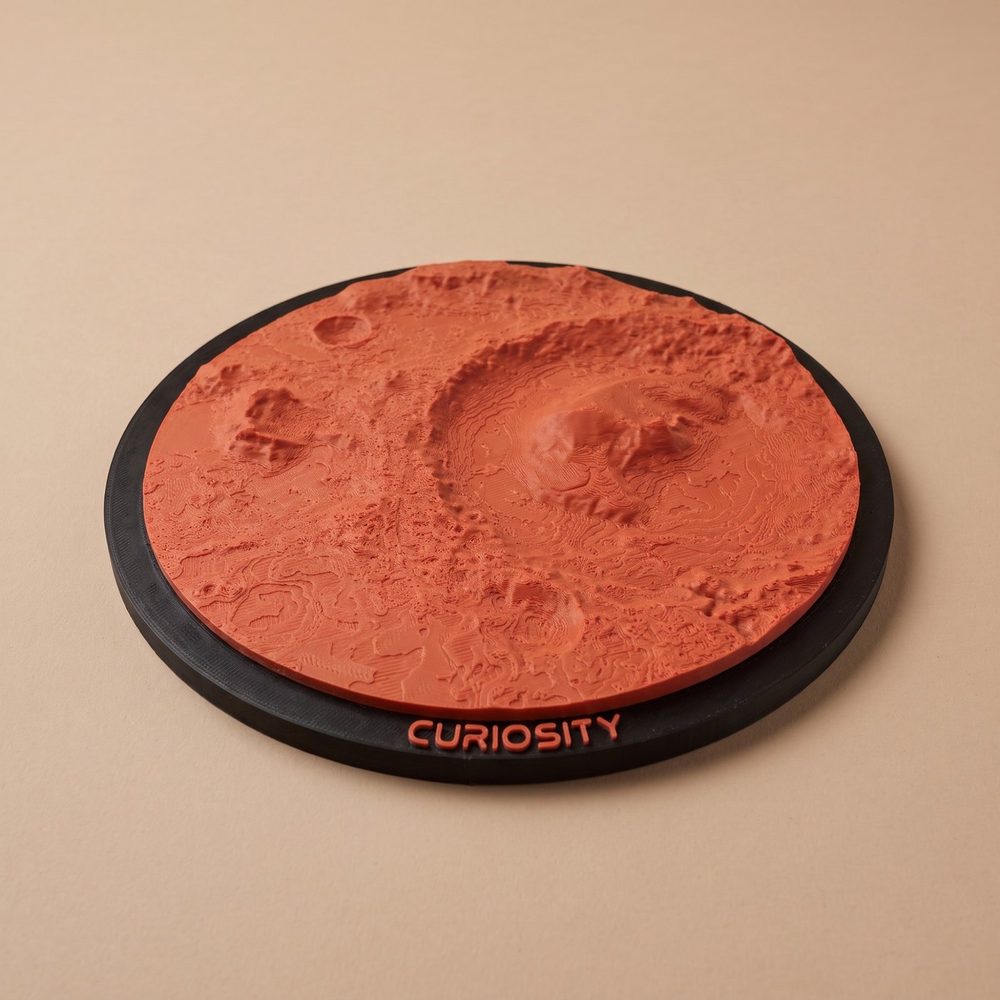 A round Mars ornament of the Curiosity landing site: Gale crater with Mount Sharp in the middle, in red on a black frame with CURIOSITY in raised letters