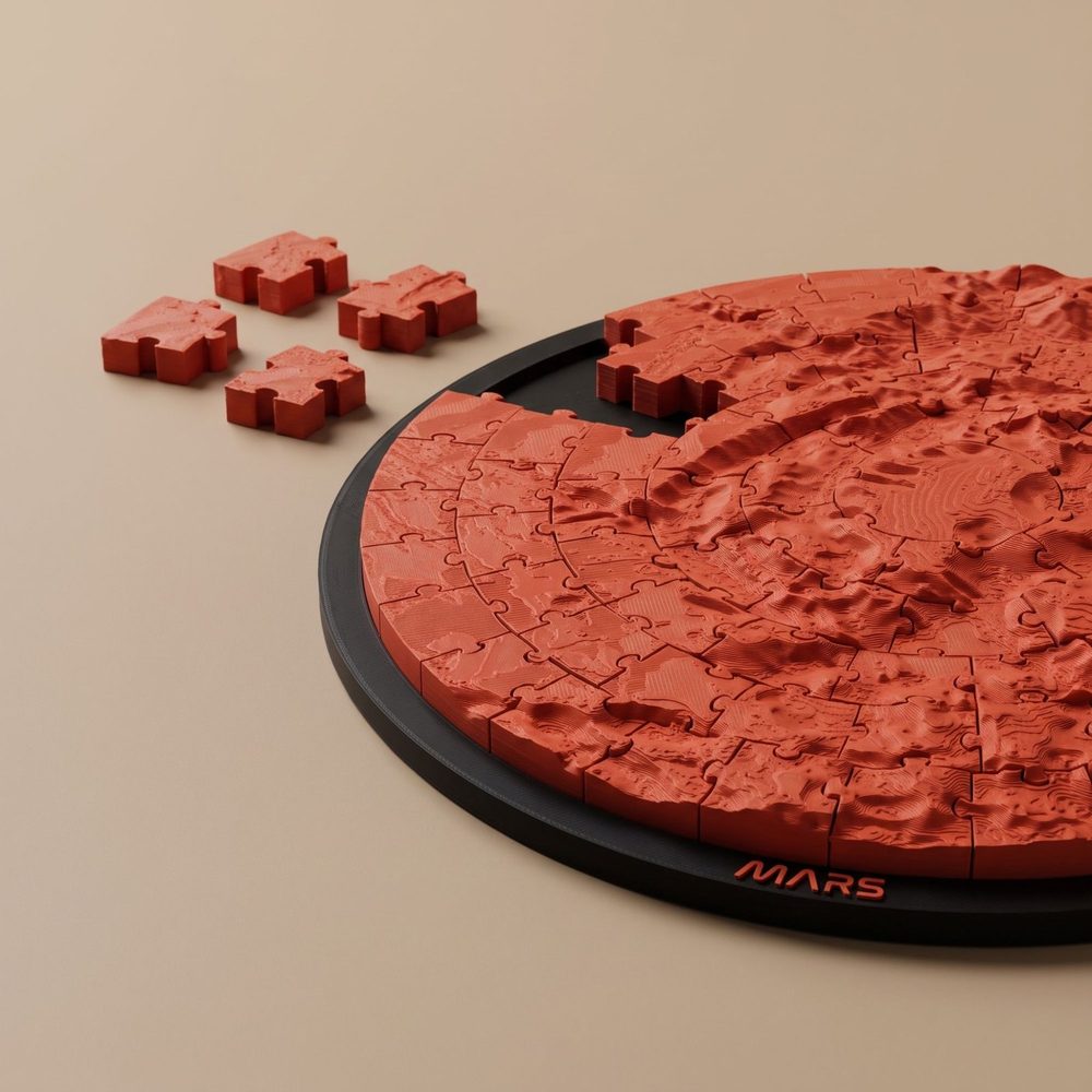 Close-up of a round Mars terrain jigsaw puzzle in red on a black frame lettered MARS, with four loose pieces beside it