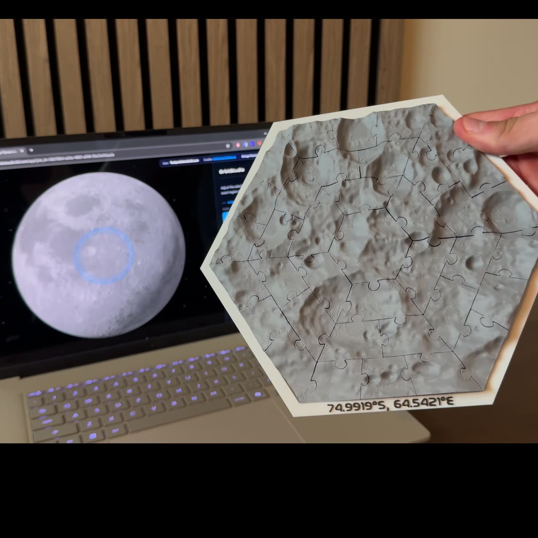 A hand holding a printed hexagon Moon jigsaw puzzle, lettered 74.9919°S, 64.5421°E, in front of a laptop running OrbitStudio